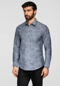 Homme aux cheveux foncés et barbe, portant une chemise ajustée à manches longues avec un motif floral subtil, associée à un pantalon noir et une montre-bracelet.