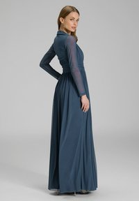 Pitkä tummansininen mekko, jossa on läpinäkyvät pitkät hihat, pleatattu vartalo-osa ja vyötäröä korostava malli, sekä virtaava helma ja sileä rakenne.