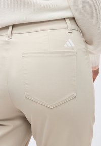 Κοντινό πλάνο πίσω τσέπης μπεζ παντελονιού με λευκό λογότυπο Adidas, φορεμένο με εκρού πλεκτό πουλόβερ.