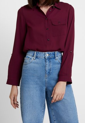 Femme portant une chemise bordeaux à manches longues avec une poche poitrine et un jean taille haute bleu clair, debout devant un fond uni.