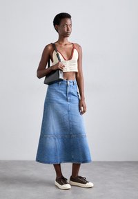 Gehaakte crop top in crème met rode accenten, gecombineerd met een lange blauwe spijkerrok. Een zwarte handtas en bruine sneakers maken de look compleet.
