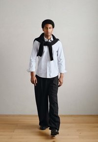 Μοντέλο που φοράει λευκό πουκάμισο με κουμπιά, φαρδιά μαύρα παντελόνια και μαύρο πουλόβερ ριγμένο στους ώμους. Λείο ύφασμα και άνετη εφαρμογή.