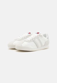 Zapatillas con una parte superior de malla y ante blanca, rayas grises, detalles texturizados y una suela de goma blanca. Discretos acentos de logo en rojo y azul.
