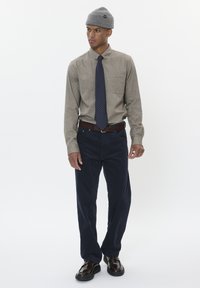 Camicia button-up beige con taschino, pantaloni di velluto a coste blu scuro, cravatta a pois navy, cintura in pelle marrone e berretto grigio in maglia, scarpe nere.