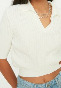 Frau in einem weißen, gerippten, kurzärmeligen, cropped Pullover mit Kragen und mehreren mit Silberketten, kombiniert mit hellblauen Jeans.