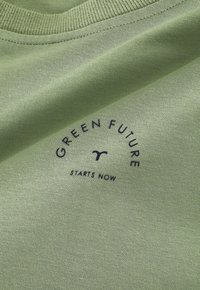 T-shirt in cotone verde con una texture morbida, con "GREEN FUTURE" in testo nero, curvato sopra, e "STARTS NOW" sotto in un design pulito.