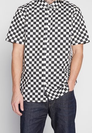 Koszula z krótkim rękawem zapinana na guziki z czarno-białym wzorem w szachownicę, noszona z ciemnymi dżinsami.