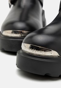 Melnas ādas potītes zābaki ar masīvu melnu zoli. Ir metāliskas akcenti tuvu purngalam, kas rāda zīmola nosaukumu sudraba burtos.