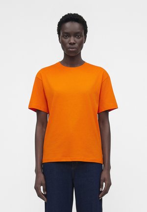 Personne aux cheveux courts et bouclés portant un t-shirt uni orange vif à manches courtes et un pantalon bleu foncé, debout devant un fond blanc.