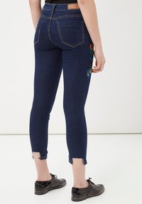 Jeans de mezclilla azul oscuro con un diseño ajustado, que presentan bordados florales en la cadera izquierda y un dobladillo desgastado. Llevados con zapatos negros.