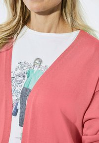Korallfarbener Cardigan mit offenem Vorderteil und strukturiertem Strick, getragen über einem weißen T-Shirt mit einer illustrierten Frau in grünem Oberteil und schwarzen Hosen.