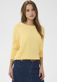Pull jaune en maille avec manches trois-quarts, associé à une jupe en denim foncé présentant un motif à rayures verticales et des poches avant.