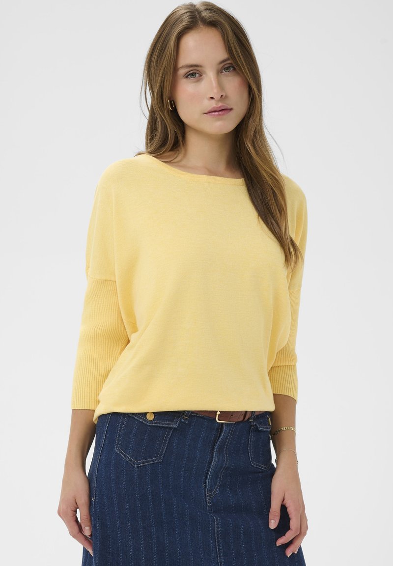 Pull jaune en maille avec manches trois-quarts, associé à une jupe en denim foncé présentant un motif à rayures verticales et des poches avant.