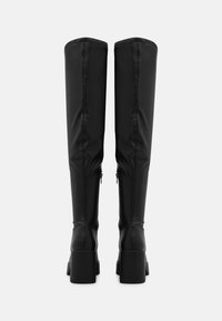 Botas negras por encima de la rodilla hechas de cuero sintético suave, con un tacón grueso, detalles de cremallera y costuras verticales en la parte trasera.