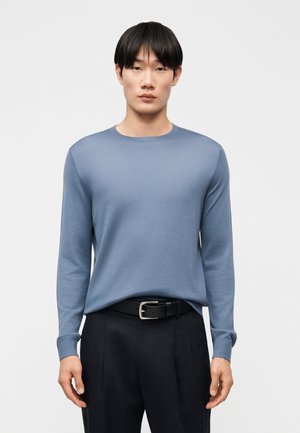 Młody mężczyzna o czarnych włosach, ubrany w jasnoniebieski sweter z długim rękawem, czarny pasek i czarne spodnie, stojący na tle jednolitego białego tła.