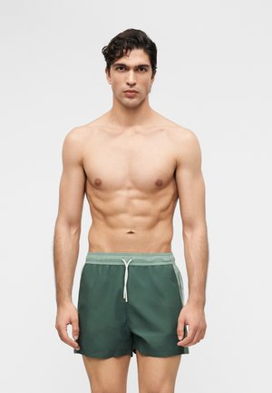 Homme athlétique aux cheveux courts, bouclés et foncés, debout face à l'avant, portant un short de bain vert avec un cordon, sur un fond clair uni.