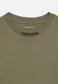 Olivenfarget T-skjorte med rund hals. Den har en jevn tekstur og brodert "Calvin Klein"-logo langs kragen.
