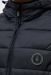 Κοντινό πλάνο μαύρου καπιτονέ μπουφάν με φερμουάρ μπροστά και κυκλικό μπαλώμα που γράφει "Jack & Jones Crafted in Blue Since 1990."