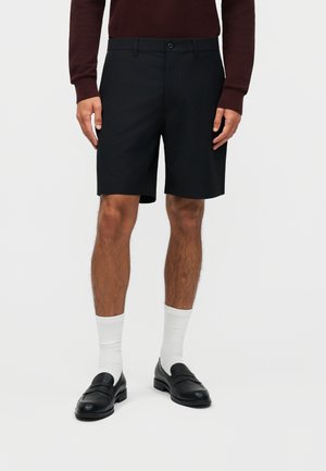 Mann trägt schwarze maßgeschneiderte Shorts, weiße Sportsocken, schwarze Ledermokassins und ein dunkles Langarmoberteil und steht vor weißem Hintergrund.