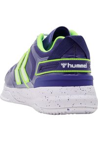 Lila idrottssko med en strukturerad ovandel, neongröna detaljer, en prickig vit mellansula och en logotyp på hälen.