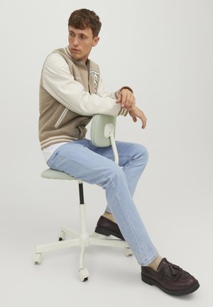 Νέος άνδρας με μπεζ varsity μπουφάν, ανοιχτό μπλε τζιν και καφέ loafers κάθεται πλαγίως σε μια λευκή καρέκλα γραφείου με ρόδες.