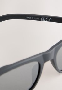 Sive sunčane naočale s mat završetkom, tamnim lećama i bočnim oznakama koje označavaju "MADE IN CHINA", "UK" i "CE".