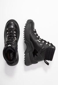 Botas de senderismo negras con acabado brillante, suela gruesa, cuello de punto texturizado y ojales de metal con cordones. Marca visible en el lateral.