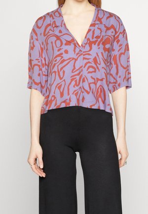Femme portant une chemise violette à manches courtes avec des motifs abstraits rouges et un pantalon noir taille haute, debout les bras détendus.