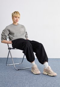 Grå fleece sweatshirt med logotyp, svarta baggy-byxor, krämfärgade sneakers. Person sittande på en metallstol mot en ljus bakgrund. Texturerad matta.