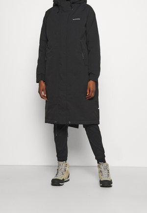 Personne portant un long manteau d’hiver noir à capuche, un pantalon noir et des bottes de randonnée beiges doublées de fourrure, debout sur un fond blanc uni.