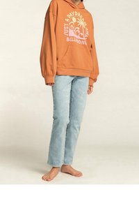 Felpa con cappuccio color arancione ruggine con testo grafico in rosa e bianco, dotata di una tasca frontale. Abbinata a jeans a gamba dritta azzurri chiari e piedi nudi.