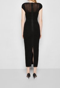 Robe noire avec des manches courtes, en tissu tricoté texturé, fermeture éclair à l'arrière et fente sur le côté. Portée avec des escarpins noirs.