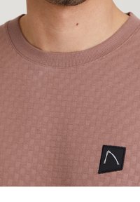 Zbliżenie na osobę w fakturowanej bluzie z okrągłym dekoltem w odcieniu mauve, z małą czarną łatą na piersi, na której znajduje się biały abstrakcyjny wzór.