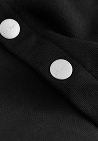 Crni materijal s teksturiranom površinom i dva srebrna gumba s ugraviranim tekstom "ALLSAINTS" za kopčanje.