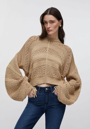 Frau trägt einen beigen, grob gestrickten Pullover mit Ballonärmeln und blauen Jeans, die Hände in den Taschen, vor einem schlichten hellen Hintergrund.