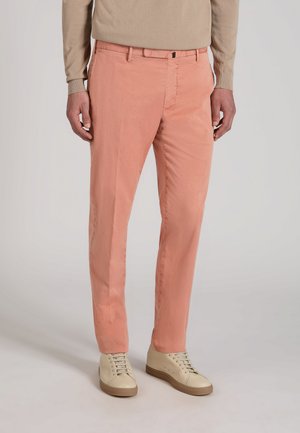 Hombre vestido con pantalones de corte recto color salmón y suéter beige, con zapatillas crema, de pie sobre un fondo claro y liso.