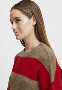 Maglione lavorato a maglia con audaci strisce rosse e beige, colletto rotondo e una morbida consistenza. Il tessuto appare caldo e accogliente.