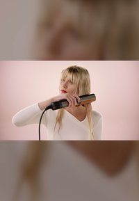 Hårplattång med en slät metallisk yta. Användaren håller den mot fuktigt hår, klädd i en räfflad vit topp mot en mjuk rosa bakgrund.