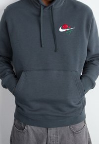 Grå hoodie i mjukt tyg, med en framficka och en broderad röd ros med gröna blad bredvid den vita logotypen på bröstet.