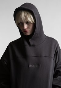 Persoon met blond haar die een donker capuchontrui draagt met het woord "wasted" geborduurd op de voorkant, kijkend naar de zijkant.