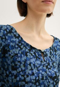 Blaue und schwarze gemusterte Bluse mit einem Rundhalsausschnitt, strukturiertem Stoff und einem kleinen Knopfverschluss am Kragen.