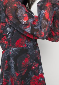 Vestido transparente con una base negra y un patrón floral en rojo y gris, mangas largas abullonadas y una cintura ajustada acentuada por un cinturón.