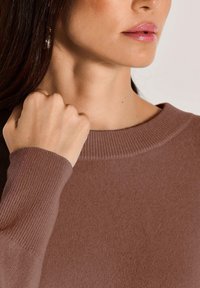 Brauner Strickpullover mit rundem Halsausschnitt, gerippten Bündchen und weicher Textur. Nahaufnahme zeigt eine Hand, die auf der Schulter ruht.