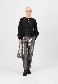 Svart blus med dekorativt band, genomskinlig textur och subtila mönster, tillsammans med metalliska silverfärgade byxor med vida ben och en silverväska. Svarta stövlar bärs.