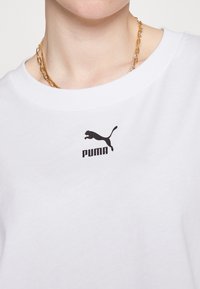 Gros plan d'une personne portant un t-shirt blanc Puma avec un logo noir et un collier chaîne en or sur un fond blanc uni.