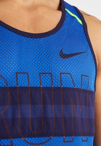 Gros plan sur une personne portant un débardeur Nike bleu perforé avec des bordures foncées et de grandes lettres partiellement visibles sur la poitrine.
