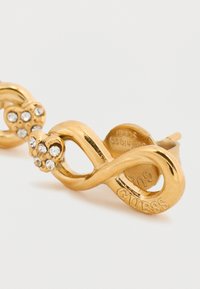 Σκουλαρίκια καρφάκια σε χρυσό τόνο, με σχέδιο άπειρου και υφή. Η μια πλευρά έχει ένα σύμπλεγμα διαφανών κρυστάλλων για επιπλέον λεπτομέρεια.