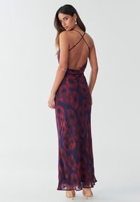 Dlouhé šaty bez rukávů z průsvitné látky s fialovým pozadím a červenými květinovými vzory, s nízkým zády a tenkými přeplétanými ramínky.