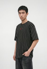 Joven con cabello negro corto, que lleva una camiseta negra de rayas finas con letras rojas y vaqueros oscuros, de pie con una mano en el bolsillo.
