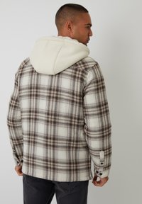 Kurtka w kratę w kolorze beżowym, brązowym i kremowym z białym kapturem wyłożonym polarem. Posiada mankiety na guziki oraz lekko teksturowany materiał.